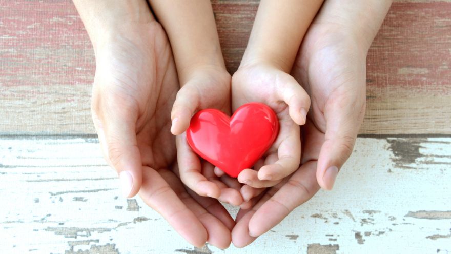 A child's hands hold a red heart while an adult's hands cup the child's hands.
