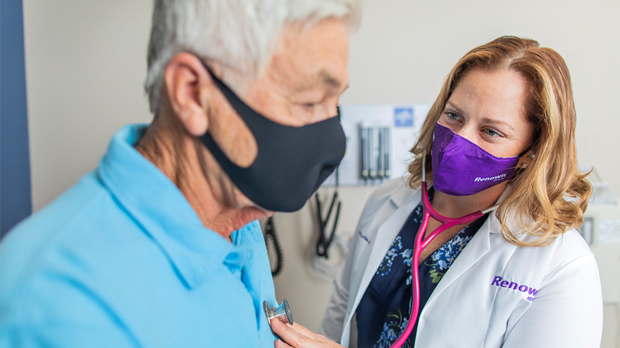 Sarah Herbert, APRN pictured wearing mask and checking masked male patient's vitals with a stethoscope 