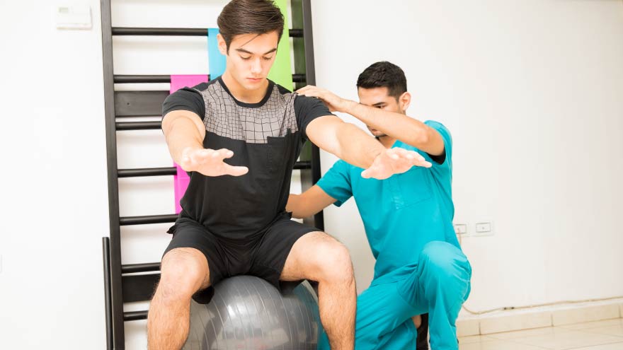Male physiotherapist helping teen balance on exercise ball